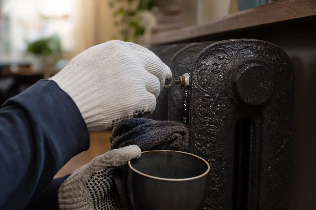 Mains gantées purgeant un radiateur en fonte avec une clé de purge, un récipient recueillant l'eau